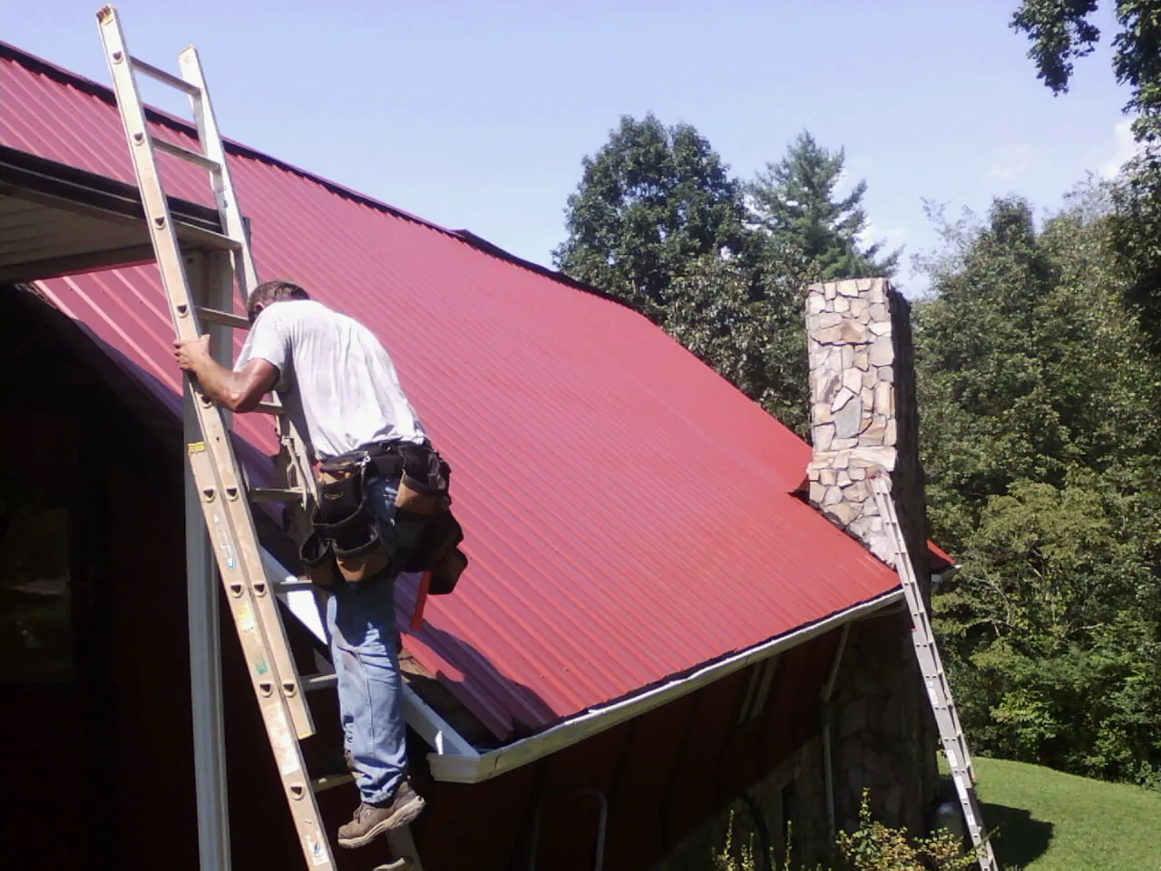 Roofer on a ladder beside a red metal roof and stone chimney — Soffit & Fascia Repair by Roofing Brothers in Winnsboro